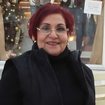 A woman with red hair and glasses standing in front of a window.